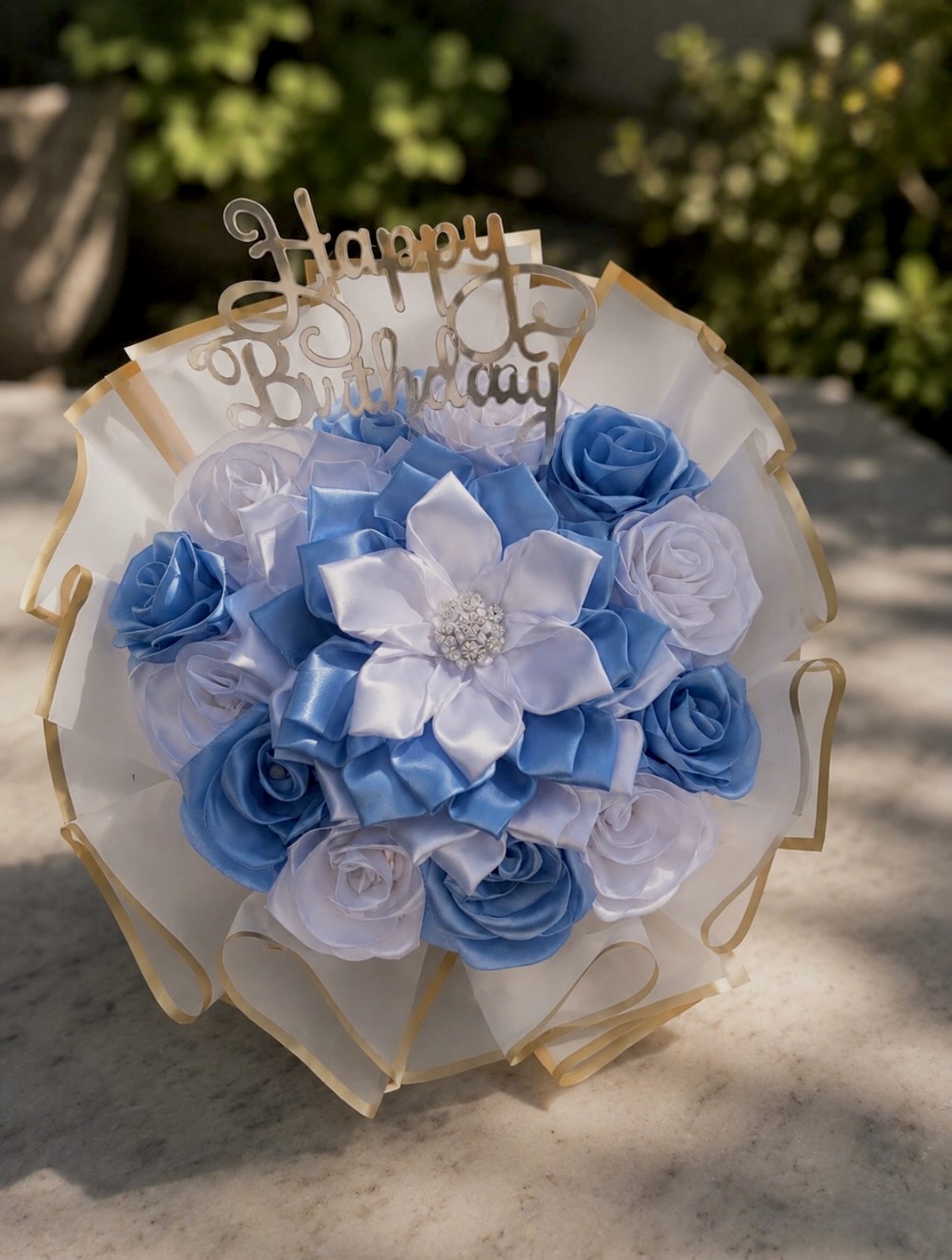 Blue and white satin ribbon rose bouquet with a pearl-centered flower and a gold Happy Birthday topper, photographed outdoors in sunlight